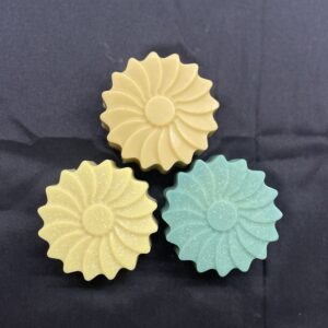 3 savons en forme de marguerite posés à plat, sur une nappe noire.