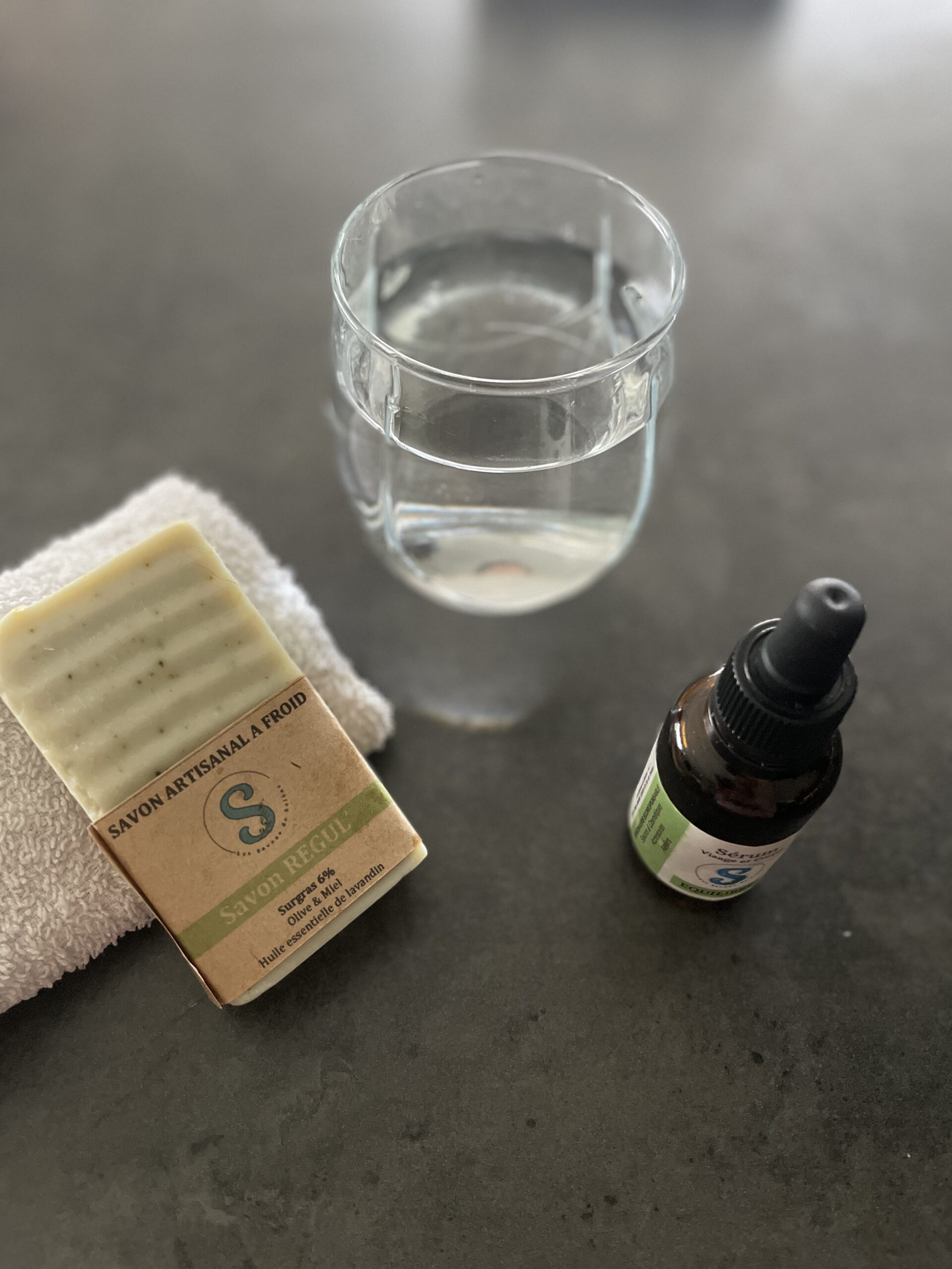 Un verre d'eau avec un flacon de sérum et un savon posé sur une serviette blanche